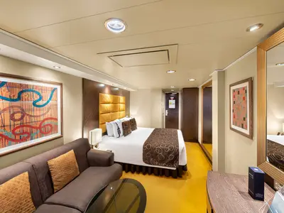 Innenansicht einer stilvollen Kabine an Bord der MSC Fantasia. Das Doppelbett, ein braunes Sofa, der gelbe Teppich und die Kunstwerke sind gut sichtbar. Helle, warme Beleuchtung.