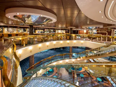 Blick in das luxuriöse, mehrstöckige Atrium der MSC Fantasia mit glänzenden Treppen, goldenen Balustraden und schicken Bars mit Lounge-Sesseln. Die Decke ist mit Lichtern geschmückt.