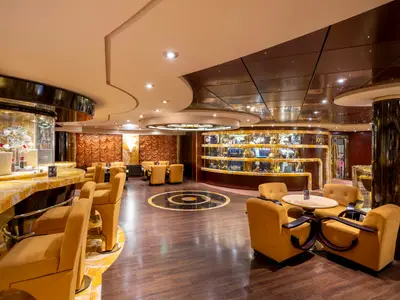 Luxuriöse Bar-Lounge 'Il Cappuccino' an Bord der MSC Fantasia. Der Raum bietet goldene Sessel, Marmortische, eine lange Theke und edle Holzvertäfelungen. Produkte in Vitrinen. Ein kreisförmiges Design ziert den dunklen Holzboden.