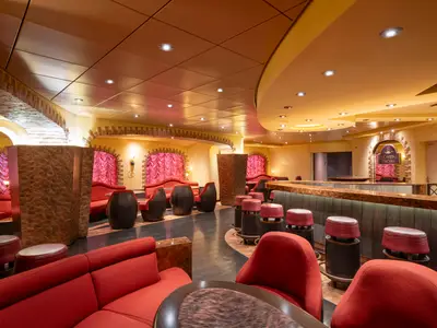 Innenansicht der La Cantina Toscana Bar an Bord der MSC Fantasia. Rote Sessel und Sofas, Holzakzente, eine lange Theke und beleuchtete Decken. Warme, einladende Atmosphaere.