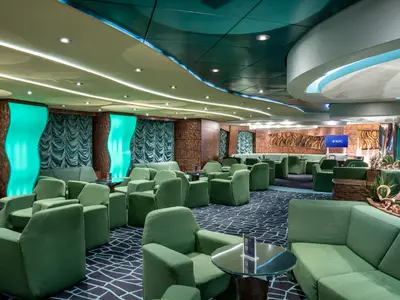 Die moderne Le Vele Bar-Lounge an Bord der MSC Fantasia zeigt gruene Sessel und Sofas, Glastische und auffaellige indirekte Beleuchtung mit aqua-farbenen Wandelementen.