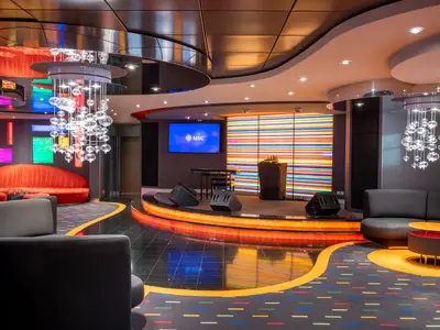 Innenansicht der modernen Manhattan Bar an Bord der MSC Fantasia. Eine Bühne mit Instrumenten und farbiger Lichtwand. Dazu elegante Leuchter und bequeme Sofas in Rot und Grau.