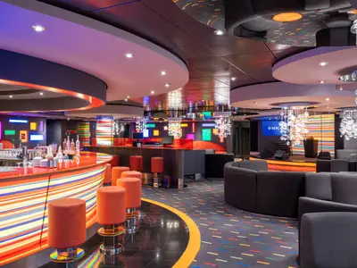 Eine farbenfrohe und moderne Manhattan Bar an Bord der MSC Fantasia, beleuchtet mit Neonlichtern. Eine bunte Bar mit Barhockern und einer kleinen Bühne ist zu sehen. Im Hintergrund sind gemütliche Sitzgelegenheiten und eine Leinwand mit dem MSC Logo.