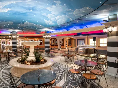 Die stilvolle Piazza San Giorgio Bar-Lounge an Bord der MSC Fantasia mit Tischen, Stühlen, zentralem Brunnen und einer Decke, die einen Sternenhimmel darstellt. Ein weißer Flügel ist im Hintergrund zu sehen.