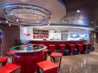 Die belebte Sports Bar an Bord der MSC Fantasia. Ein langer, roter Tresen mit passenden Barhockern. Im Hintergrund Bildschirme und Regale voller Flaschen.