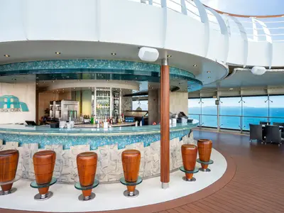 Blick auf die Bar Delle Fontane an Bord der MSC Fantasia. Eine geschwungene Bar mit Holzstuehlen und blauen Mosaikfliesen, umgeben von Panoramafenstern mit weitem Meerblick.