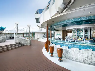 Die Bar Delle Fontane an Bord der MSC Fantasia. Eine geschwungene Outdoor-Bar mit blauen Mosaiken, Holzhockern und Getränken auf dem sonnigen Deck. Moderne Skulpturen sind sichtbar.