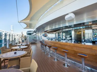 Outdoor-Bar, die Gaudi Bar, an Bord der MSC Fantasia. Mit langer Holztheke, Hockern und Rattan-Sitzgruppen unter einem Sonnensegel. Blick auf Meer und Hafen.