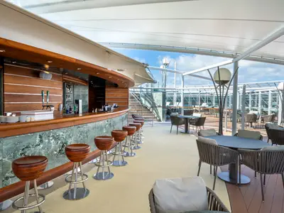 Outdoor-Bar The One Bar auf dem Sonnendeck der MSC Fantasia. Holztheke, Barhocker, Lounge-Sitzgruppen und Tische unter einem Sonnensegel. Blauer Himmel.
