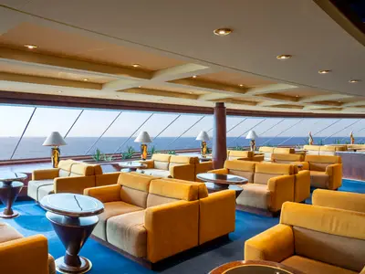 Elegante Top Sail Lounge an Bord der MSC Fantasia. Gemütliche gelbe Sofas, Tische und große Fenster mit Panoramablick auf das weite Meer. Moderne Beleuchtung schafft eine luxuriöse, entspannte Atmosphäre.