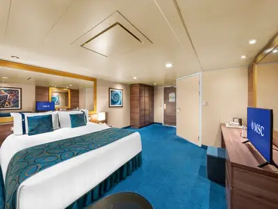 Geräumige barrierefreie Innenkabine an Bord der MSC Fantasia. Sichtbar sind ein großes Doppelbett mit türkiser Tagesdecke, ein Schreibtisch mit TV und ein Kleiderschrank. Blauer Teppichboden.