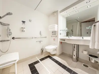 Ein modernes, barrierefreies Badezimmer an Bord der MSC Fantasia mit weissen Fliesen, Duschbereich inklusive Klappsitz und Haltegriffen, einer Toilette und einem Waschtisch mit Spiegel.