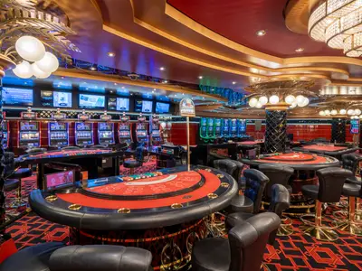 Blick in das elegant beleuchtete Casino Delle Palme an Bord der MSC Fantasia. Mehrere Spieltische mit roten Filzflächen und schwarzem Leder sind im Vordergrund und Mittelgrund zu sehen, umgeben von gepolsterten Barhockern. Im Hintergrund stehen Reihen von leuchtenden Spielautomaten. Die Decke ist rot und goldfarben gestaltet, und große Kronleuchter spenden warmes Licht. Der Teppich hat ein modernes geometrisches Muster in Rot und Schwarz.