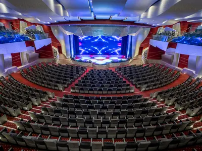Blick auf das leere Theater 'Teatro L'Avanguardia' der MSC Fantasia. Roter Teppich und Reihen grauer Sessel führen zur hell beleuchteten Bühne mit blauer Sternenprojektion.