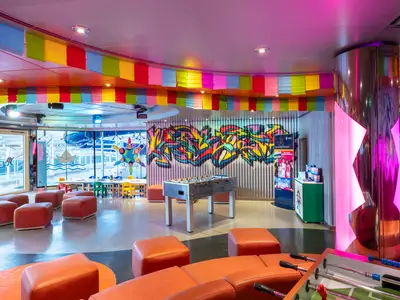 Ein lebhafter Kinder- und Teenagerbereich an Bord der MSC Fantasia. Mit Tischkicker, bunten Sitzpoufs, Graffiti und pink beleuchteter Säule.
