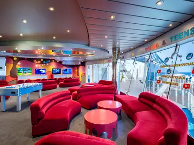 Der Young & Teen Club an Bord der MSC Fantasia ist ein bunter Raum mit roten Sofas, einem Foosballtisch und Bildschirmen für Videospiele. Große Fenster bieten Meerblick.