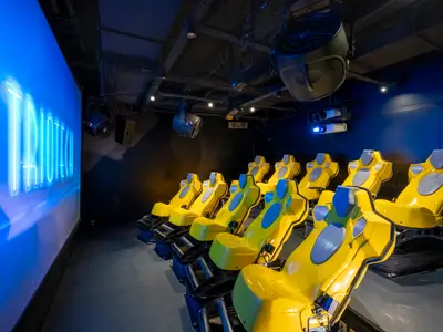 Ein 4D-Kino an Bord der MSC Fantasia mit leuchtend gelben Sitzen und blauem Bildschirm mit 'TRIOTECH'-Schriftzug. Dunkle Wände und Decke mit Technik.
