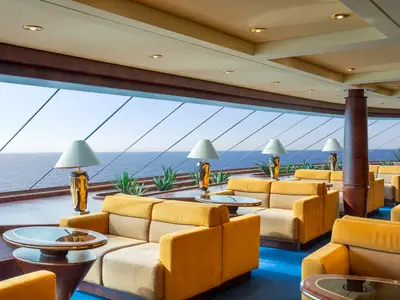 Elegante Lounge an Bord der MSC Fantasia mit großen Panoramafenstern und weitem Meerblick. Bequeme gelbe Sofas und Tische, beleuchtet von modernen Stehlampen und Pflanzen.