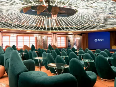 Innenansicht der L'Insolito Lounge an Bord der MSC Fantasia, featuring tealfarbene Sessel, runde Glastische und eine große Leinwand mit dem MSC Logo. Die Decke ist mit einer auffälligen Lichtinstallation verziert.