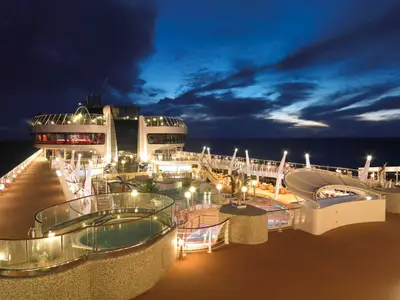 Blick auf das beleuchtete Pool- und Sonnendeck an Bord der MSC Fantasia bei Nacht. Mehrere Pools, Jacuzzis und eine Lounge sind sichtbar, unter einem dunklen Nachthimmel mit Meerblick.