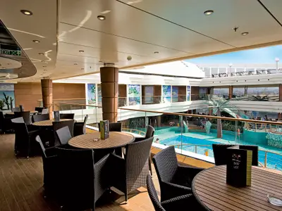 Das Pool-Atrium der MSC Fantasia. Im Vordergrund Tische und Korbsessel einer Bar. Dahinter ein mehrstöckiger Poolbereich mit Palmen, Wasserfontänen und Mosaikfliesen. Mehrere Personen sichtbar.