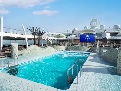 Der sonnige Aquapark an Bord der MSC Fantasia. Man sieht einen großen Pool mit Wasserfontänen, Liegestühle und eine Videoleinwand unter blauem Himmel.