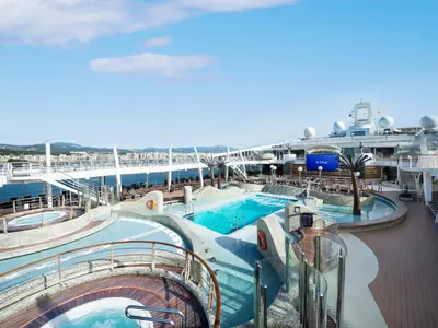 Blick auf den weitläufigen Aquapark der MSC Fantasia mit mehreren Pools, Whirlpools und Liegeflächen unter blauem Himmel. Im Hintergrund ist eine Küstenstadt zu sehen.