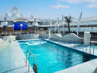 Der Aquapark an Bord der MSC Fantasia. Grosser Pool mit Wasserfällen, Fontänen und blauen Mosaikfliesen. Im Hintergrund: Sonnendecks, MSC-Leinwand und Palme unter blauem Himmel.