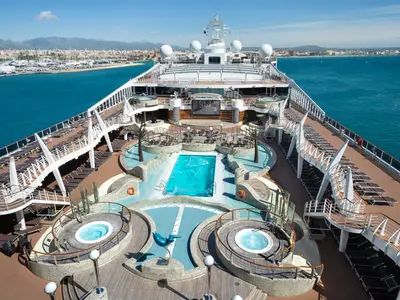 Blick von oben auf das Sonnendeck der MSC Fantasia mit dem Aquapark. Mehrere Pools, Whirlpools, Sonnenliegen und Palmen sind sichtbar. Im Hintergrund eine Küstenstadt am Meer.