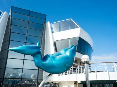 Glänzende, türkisblaue Skulptur auf dem Sonnendeck der MSC Fantasia. Moderne Glasarchitektur des Schiffes spiegelt blauen Himmel. Eine weiße Reling ist rechts zu sehen.
