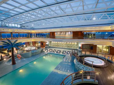 Der mehrgeschossige Innenpoolbereich 'I Tropici' an Bord der MSC Fantasia bei Abendstimmung. Ein grosser Pool, Whirlpools und Palmen sind unter einem Glasdach sichtbar. Viele Lichter erzeugen eine warme Atmosphäre.