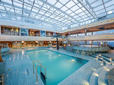 Der lichtdurchflutete Innenpoolbereich "I Tropici" an Bord der MSC Fantasia zeigt ein großes Schwimmbecken unter einem Glasdach. Palmen, Mosaike und Sitzbereiche schaffen tropisches Ambiente.