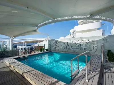 Der The One Pool an Bord der MSC Fantasia. Heller blauer Pool, umgeben von Holzdecks und einer weissen Überdachung. Im Hintergrund eine graue Wand mit abstraktem Wandrelief. Blauer Himmel.