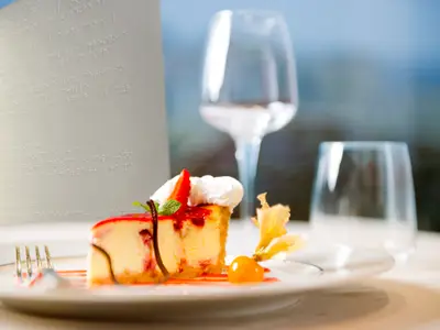 Ein Stück Käsekuchen mit Erdbeere und Sahne auf einem Teller, daneben ein Braille-Menü und zwei Gläser, an Bord der MSC Fantasia.