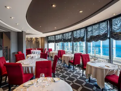 Ein elegantes Restaurant an Bord der MSC Fantasia. Mehrere Tische sind mit weissen Tischdecken gedeckt und von roten Stühlen umgeben. Grosse Fensterfronten bieten einen weiten Blick auf das offene Meer.