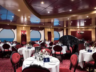 Das stilvolle Restaurant an Bord der MSC Fantasia zeigt festlich gedeckte Tische mit roten Samtstühlen und große Bullaugen mit Blick auf das blaue Meer.