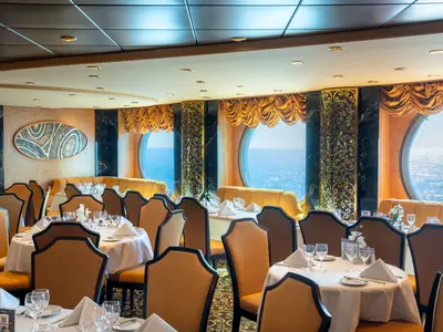 Eleganter Speisesaal der MSC Fantasia. Gedeckte Tische, braune Stühle und große Bullaugen mit Meerblick dominieren den Raum. Goldene Akzente und dunkler Marmor prägen das Interieur.