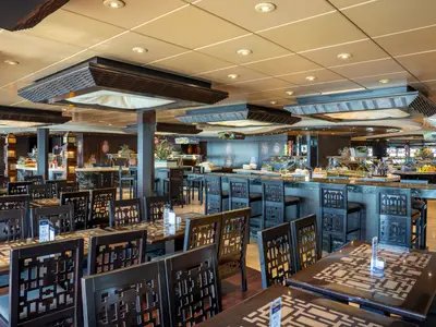 Das L'Africana & Zanzibar Buffet an Bord der MSC Fantasia. Ein grosser Speisesaal mit vielen dunklen Holztischen und Stühlen im Vordergrund. Im Hintergrund befinden sich mehrere Buffetstationen mit Speisen. Die Decke ist hell und mit dekorativen Holzelementen gestaltet, die eine warme Atmosphäre schaffen.