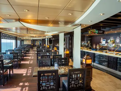 Das Buffetrestaurant L'Africana & Zanzibar der MSC Fantasia mit Meerblick. Reihen dunkler Tische und Stühle. Buffetbereich mit Speisen und afrikanischen Masken an der Wand.