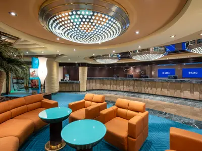 Das Bild zeigt die elegante Rezeption und eine angrenzende Lounge an Bord der MSC Fantasia. Eine gebogene Theke, blaue Bildschirme mit MSC Logo, orangefarbene Sofas und Sessel sowie eine Palme sind zu sehen. Grosse, runde Kronleuchter schmuecken die Decke.