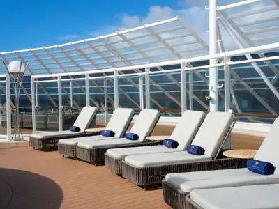 Ein Bereich an Bord der MSC Fantasia mit bequemen Sonnenliegen, blauen Handtüchern und einem modernen Lichtobjekt unter einer Glasdachkonstruktion. Blauer Himmel ist sichtbar.