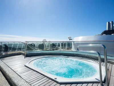 Ein sprudelnder Whirlpool auf dem sonnigen Oberdeck der MSC Fantasia. Holzplanken und Glasgeländer umgeben den Pool, im Hintergrund sind Liegen und das Meer zu sehen. Der Himmel ist blau.