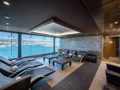 Ein moderner Spa-Ruhebereich an Bord der MSC Fantasia mit Liegen und einem beeindruckenden Panoramafenster, das den Blick auf das Meer und eine Küstenstadt freigibt. Eine Steinfassade und gedämpfte Beleuchtung schaffen eine entspannte Atmosphäre.