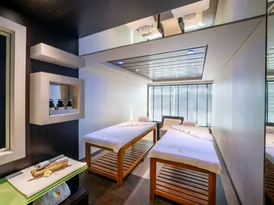 Doppel-Massageraum im MSC Aurea Spa an Bord der MSC Fantasia. Zwei hölzerne Behandlungsliegen mit weissen Handtüchern, Sessel, Fenster mit Jalousien. Links Regale mit Spa-Produkten und Bambus-Deko.