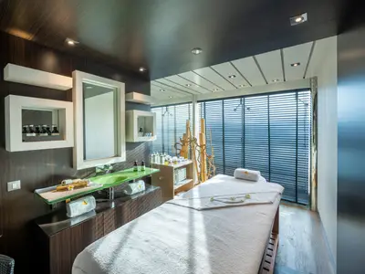 Heller Spa-Behandlungsraum an Bord der MSC Fantasia mit Massageliege, Waschtisch und großem Fenster mit Jalousien. Tageslicht für Wellness-Anwendungen.