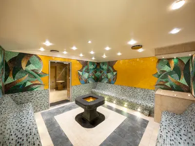 Der Thermalbereich im MSC Aurea Spa an Bord der MSC Fantasia mit bunten Mosaikwänden in Gelb und Grün. Beheizte Sitzbänke und ein zentrales Mosaikbecken sind zu sehen. Eine Glastür führt zu den Duschen.
