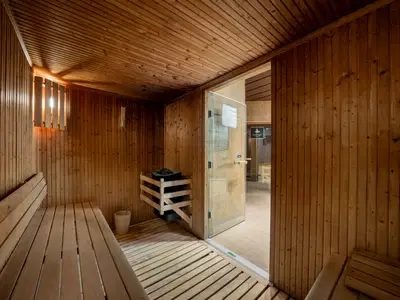 Eine holzgetäfelte Sauna im MSC Aurea Spa an Bord der MSC Fantasia, mit hellen Sitzbänken, einem Saunaofen hinter einem Holzgitter und einer hölzernen Schöpfkelle. Helles Licht fällt durch eine vertikale Holzlamellenabdeckung. Eine teilweise geöffnete Glastür führt in einen gefliesten Bereich.