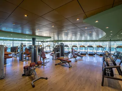 Geräumiges Fitnessstudio an Bord der MSC Fantasia mit Panoramablick auf das Meer. Moderne Technogym-Geräte, darunter Laufbänder, Kraftstationen und Hanteln, stehen auf hellem Holzboden bereit.