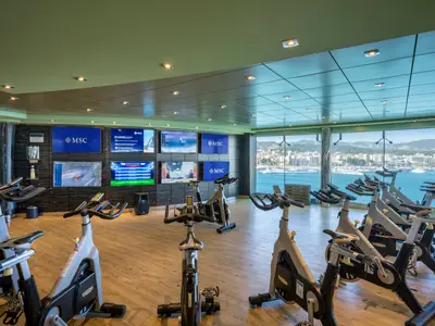 Fitnessbereich der MSC Fantasia mit Technogym-Ergometern. Große Panoramafenster zeigen eine Hafenstadt mit Yachten und Bergen. Mehrere Bildschirme an den Wänden. Modern und hell.
