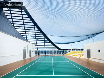 Der Multifunktions-Sportplatz an Bord der MSC Fantasia mit grünem Spielfeld, Basketballkorb und gelben Sitzen. Ein Netz schützt nach oben. Im Hintergrund sind das Meer und der blaue Himmel sichtbar. Keine Personen.